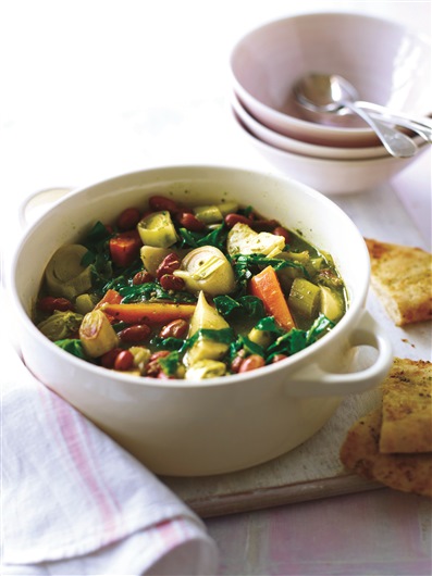 This image shows a bowl of spring vegetable casserole. The vegetables are cut into chunks and are brightly coloured. Next to the bowl are some slices of garlic bread.