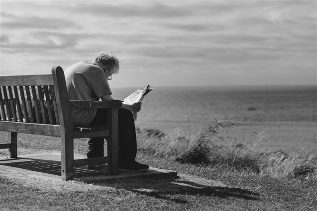  Black and white photograph of a man on a bench beside the seafront, facing away from the camera and looking down at a newspaper