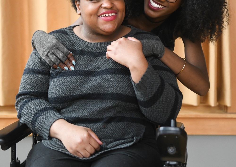 Close-up of a couple smiling and holding hands. The Black femme in the back wears compression gloves and looks lovingly at the non-binary Black person in the front, who sits in a power wheelchair.