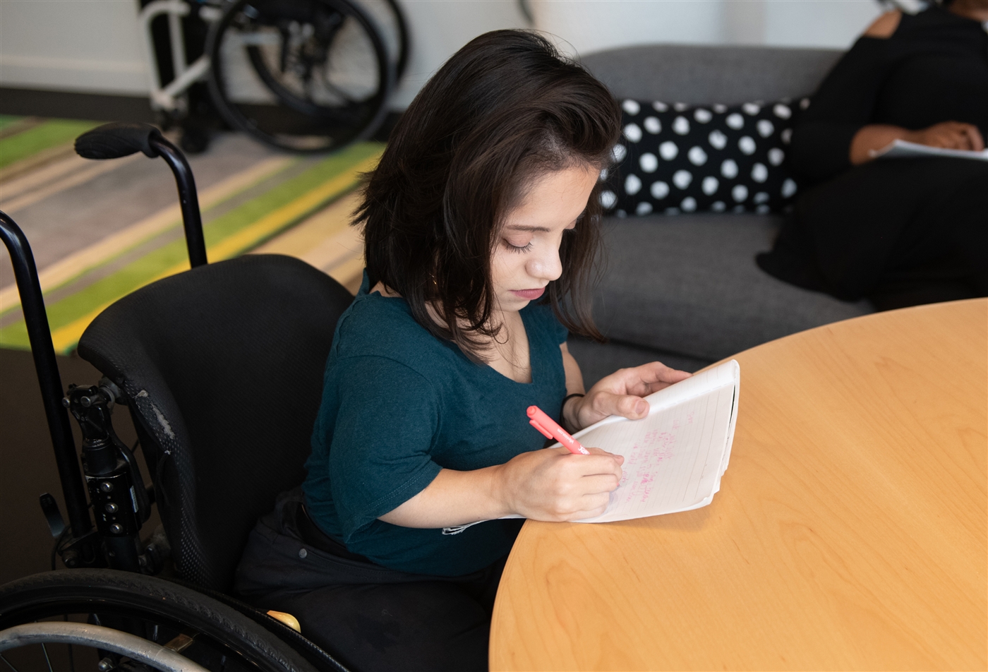 A South Asian person in her wheelchair takes notes by hand during a meeting.