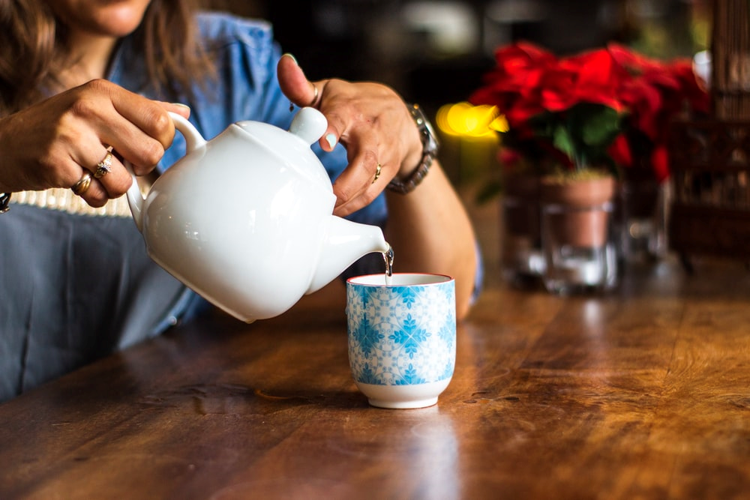 A woman pouring tea into a blue patterned teacup on a rustic wooden table.
