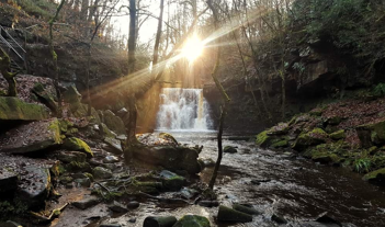 A photo of a waterfall in woodland with low sun shining through the trees