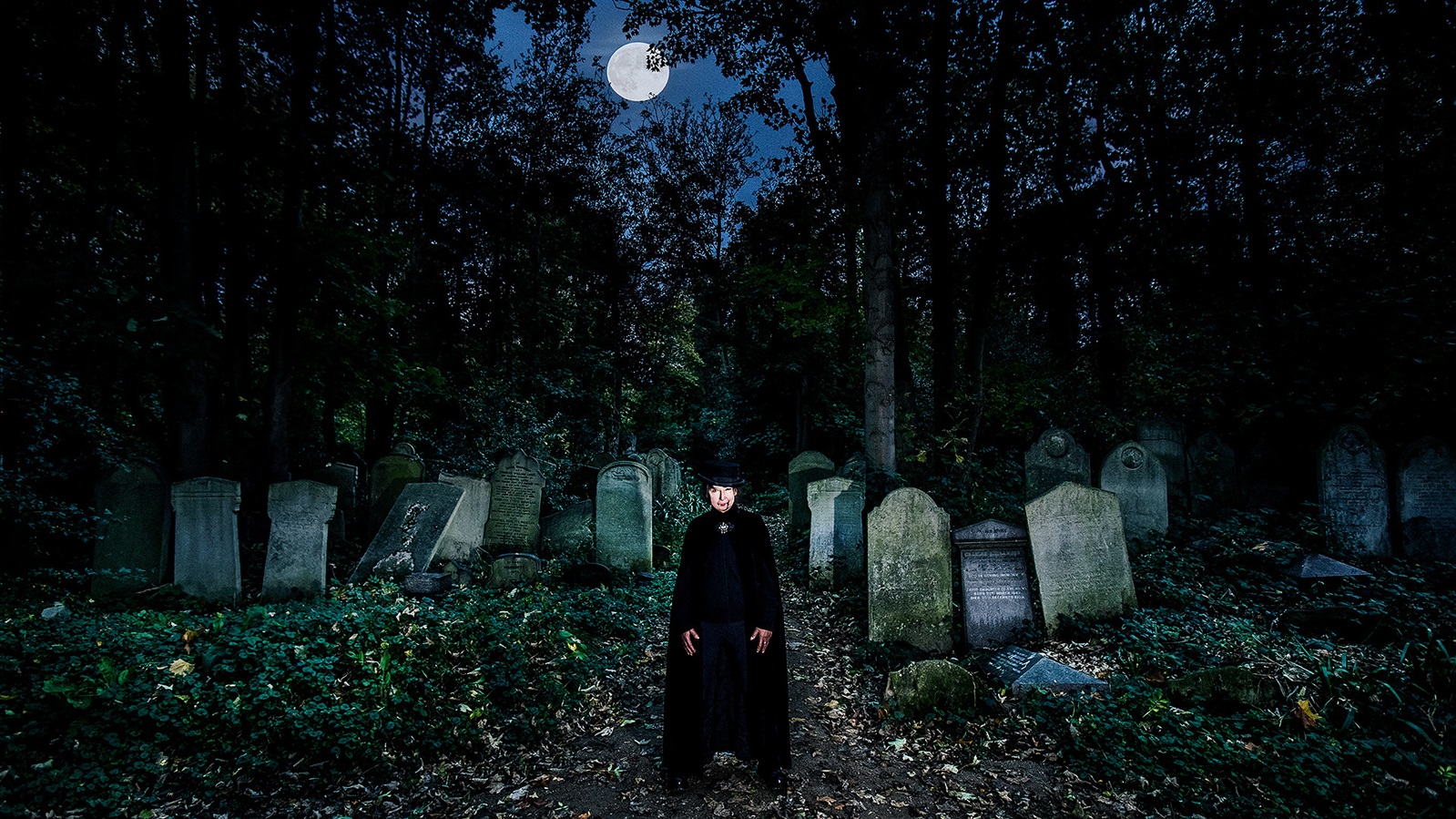 Woman in long cloak standing amongst gravestones in a cemetery with a full moon overhead.
