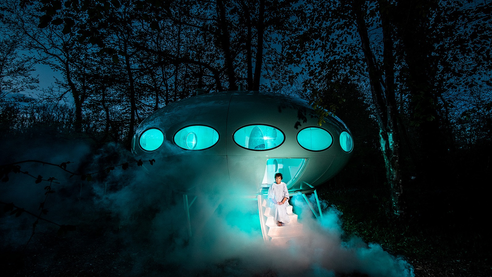 Biopsy. Woman in hospital gown sits on the steps of a spaceship in a forest at night.