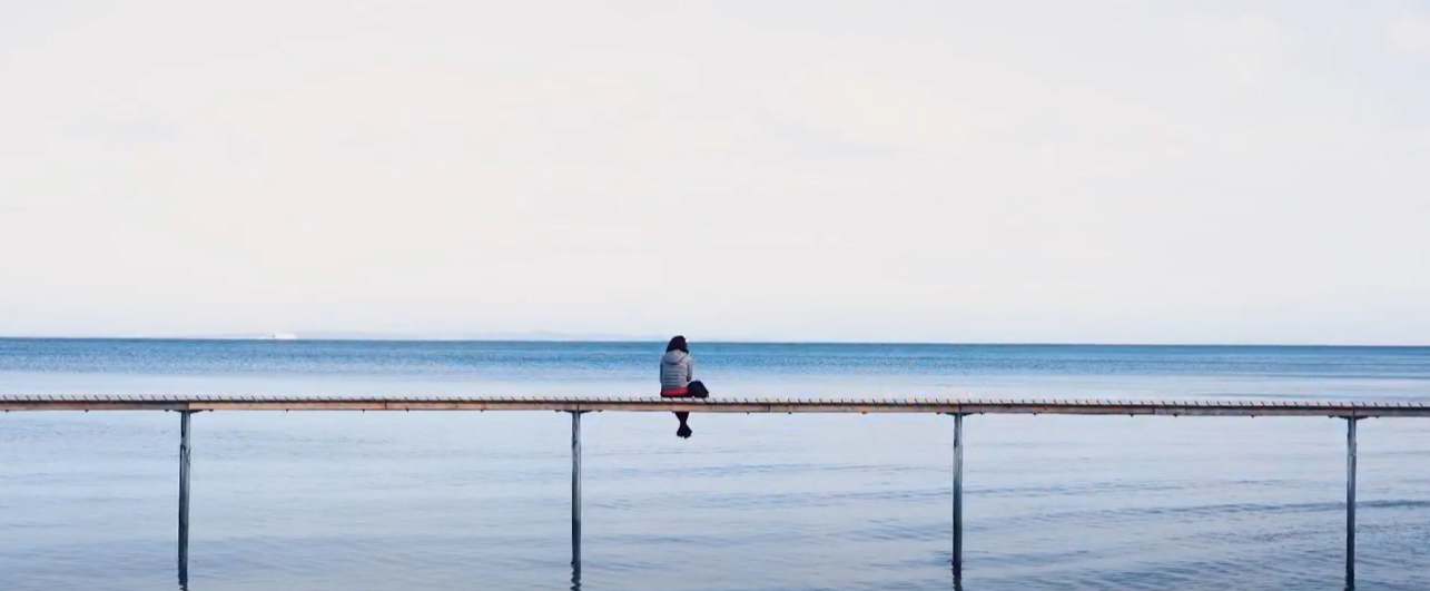 A woman sits alone on a pier and looks out to sea. The sky is pink and yelllow like a sunrise or sunset.