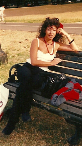 Willo sits on a bench in London on a summers day looking into a camera with a red flower in her hair. 