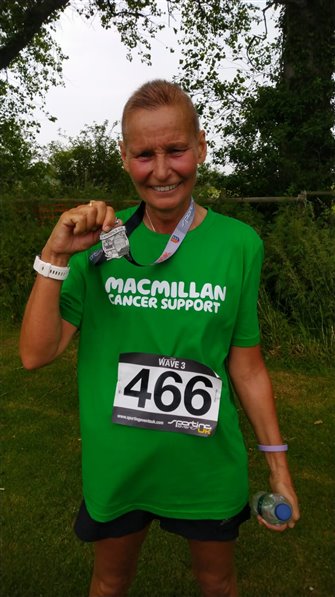 Linda is smiling and holding up her medal for running 10k, while wearing her green Macmillan t shirt.