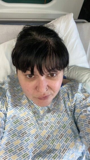 Katy has brown hair and brown eyes and a full fringe. She is lying on a bed in a hospital gown and taking a selfie.