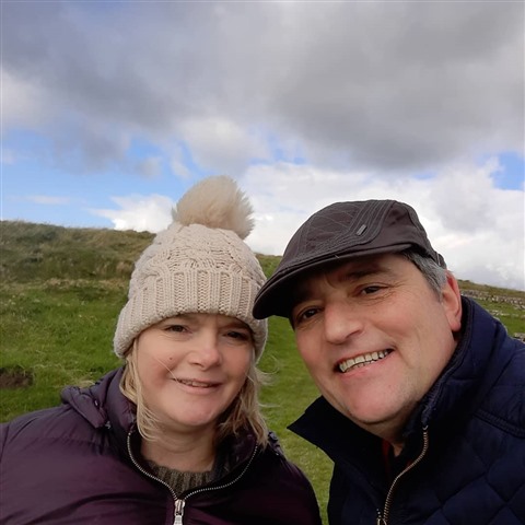 A photo of John and a family member on a walk in the countryside, both are smiling at the camera and wearing warm hats.