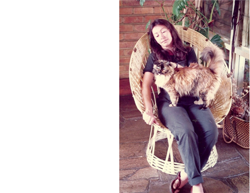  A woman sat on a wicker chair with a cat on her lap outside with brickwork behind her.