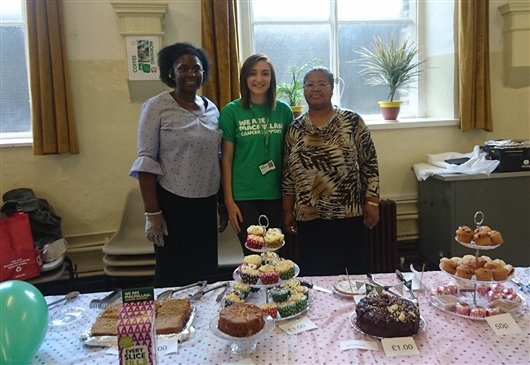 This photo shows three smiling women, standing behind a table full of cakes. The woman in the centre is wearing a green Macmillan T-shirt, and the other two are dressed normally.