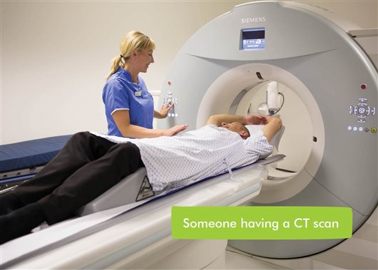 This image shows someone lying down to have a CT scan. There is a health professional assisting them.