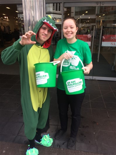 This photo shows two smiling people outside an M&S store, holding Macmillan fundraising buckets. On the left is a man, wearing a green animal onesie, and on the right is a woman wearing a green Macmillan T-shirt. 