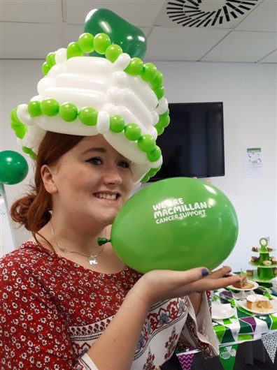 This photo shows a smiling woman wearing a hat made of green and white balloons. She is holding a green balloon with the Macmillan logo on it. Behind her is a table full of cakes.
