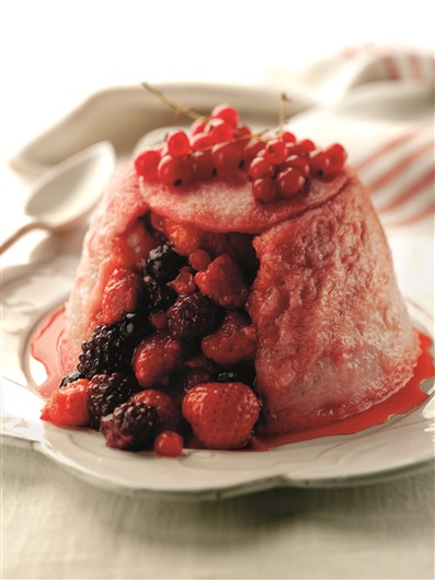 This image shows a summer pudding that has been cut into. The pudding is topped with berries, and the filling of berries is spilling om the berry juut of the cut. The sponge is slightly pink in colour from the berry juice.