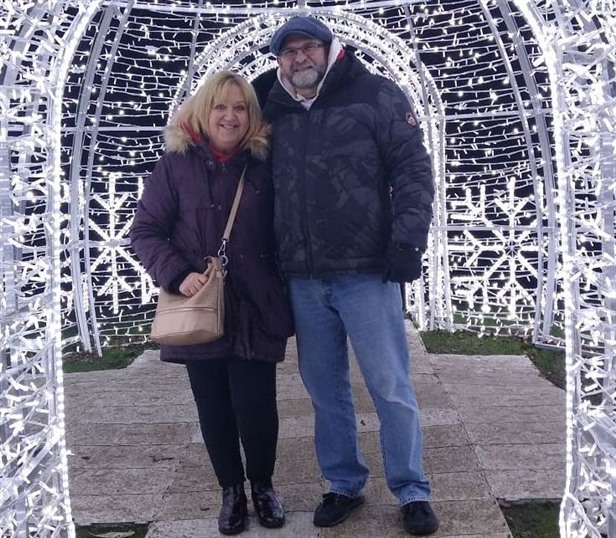  A photograph of a man and a woman, with Christmas lights in the background. The man has his arm around the woman, and they are both smiling at the camera and wearing warm winter clothes.