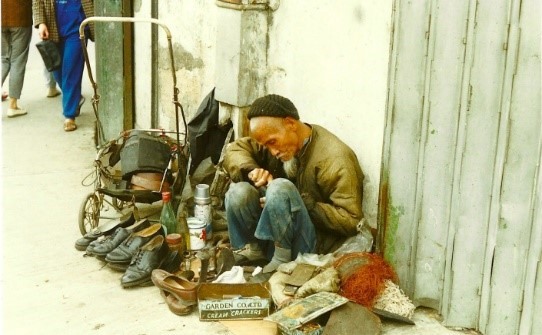 A photograph taken by Willo of a cobbler mending shoes. The cobbler is sitting and working on a shoe and surrounded by pairs of finished shoes.