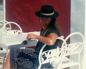 A photograph of Willo, sitting outside at a white table and chairs smiling into the camera. She is wearing a black sunhat and holding sunglasses.