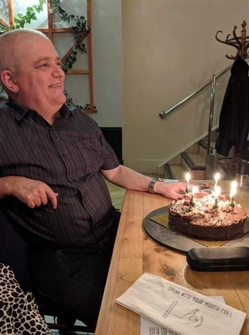 Photo of John sitting at a table with a chocolate birthday cake in front of him candles lit. 