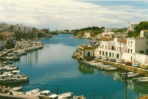 A photograph of a sunny river in Menorca from a high vantage point, there are white fishing boats on the water and lovely house on the banks.