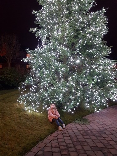 A little girl in a pink coat, sat next to a giant pine tree covered in Christmas lights.