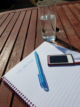 A sunny outdoor table with a glass of water, notepad and a pen.